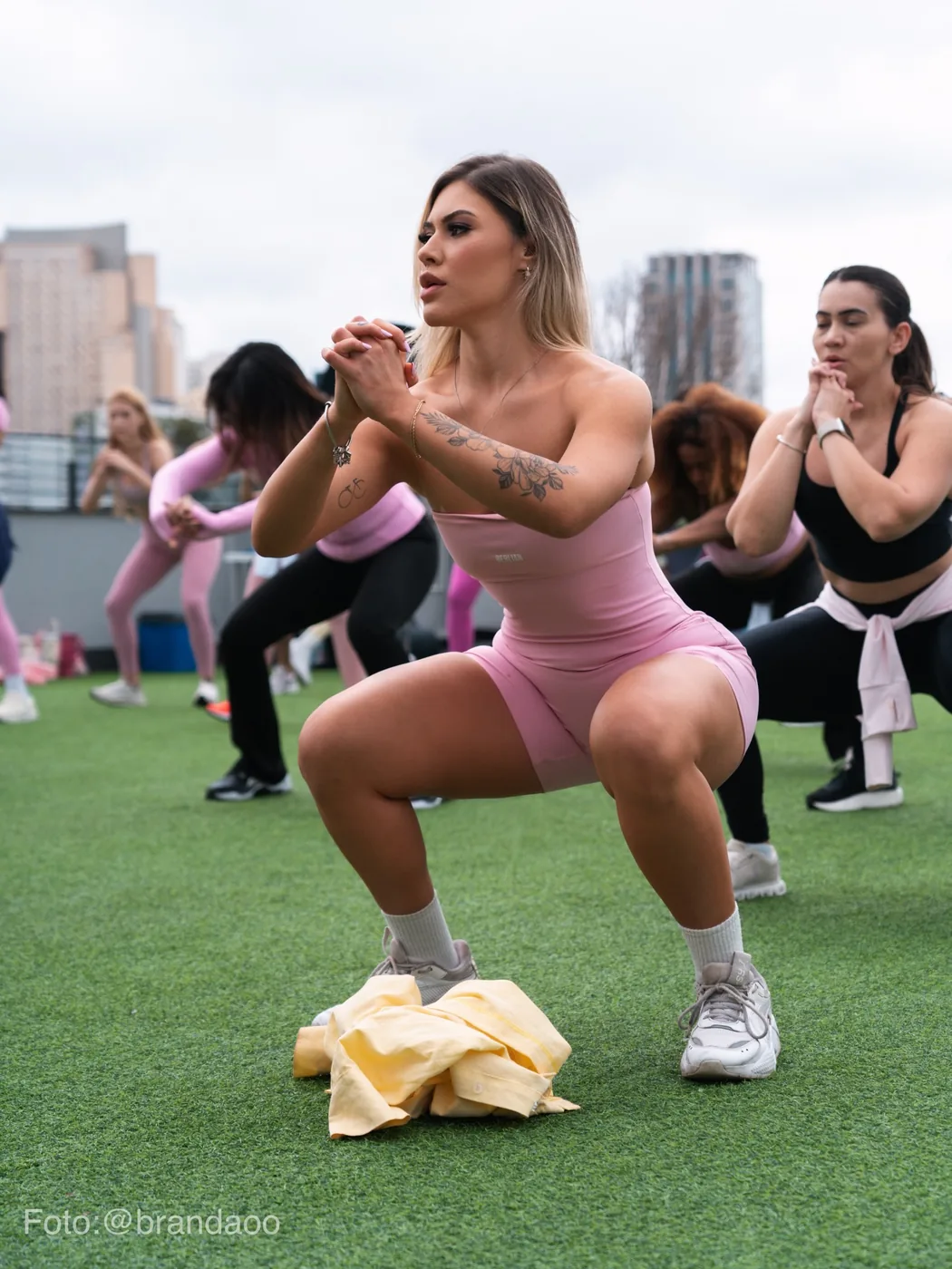 Bre Cardoso agachada no gramado do rooftop, treinando com participantes da primeira edição