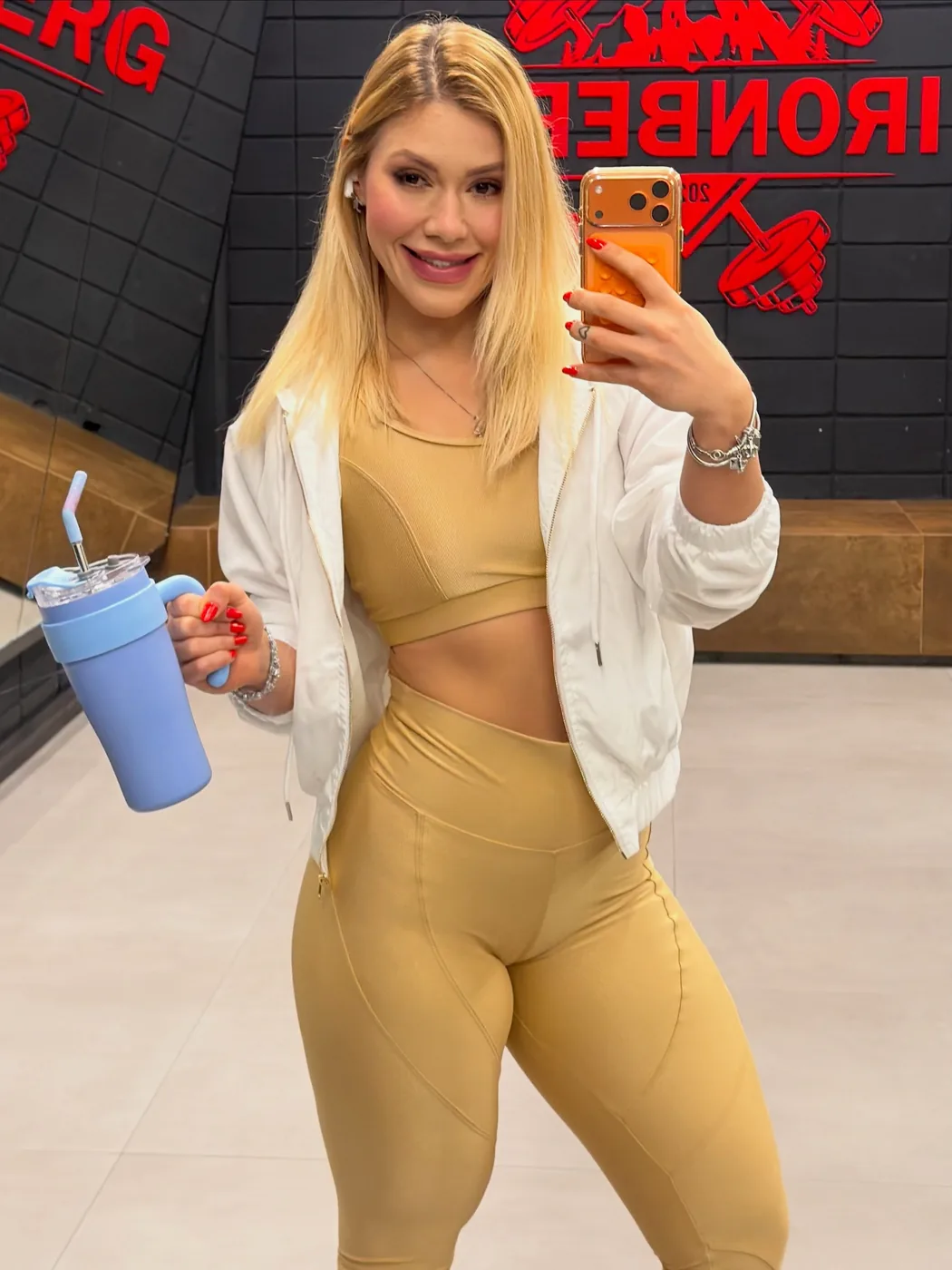 Bre Cardoso de conjunto dourado de treino e jaqueta branca, selfie na academia